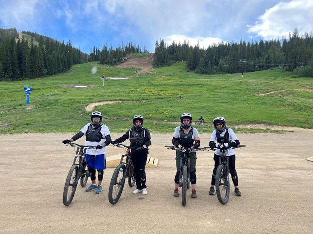 Four mountain bikers stand at the foot of a trail they just biked down.